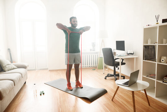 Healthy life. Full length shot of male fitness instructor showing exercises with resistance band while streaming, broadcasting video lesson on training at home using laptop