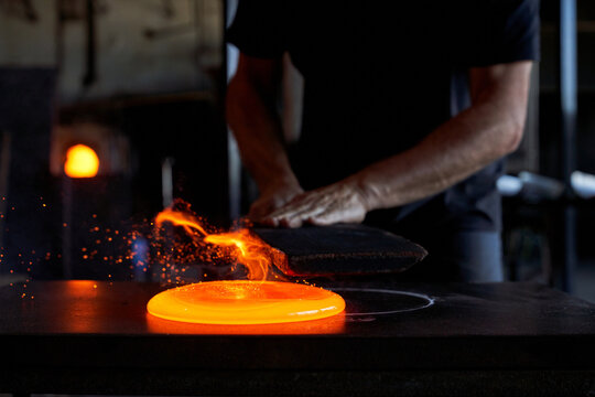 Faceless man working with liquid hot glass while blowing art elements in dark workshop