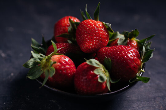 Closeup of delectable juicy ripe strawberries with green leaves arranged in black ceramic bowl on dark background