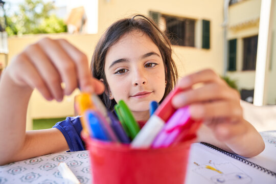 Positive school aged girl while sitting at table and choose drawing with colorful pencils in copybook during free time