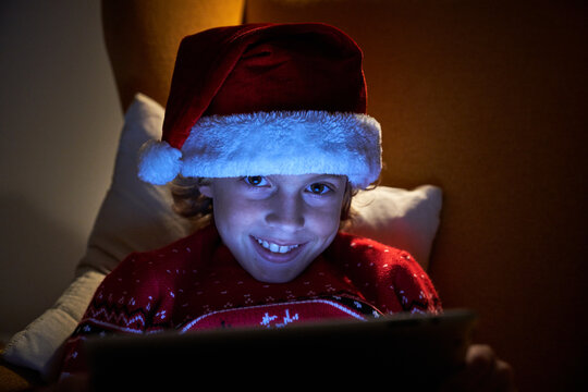 Cheerful Boy In Christmas Wear Using Social Media On Tablet While Sitting On Chair During New Years Holiday In Apartment