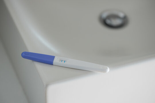 From above of pregnancy test with two bars for positive result placed on edge of white sink in bathroom