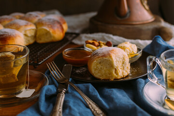 From above of ceramic plate with fresh bun and small bowls with mix of nuts near jam and butter close to cup of green tea on crumpled towel