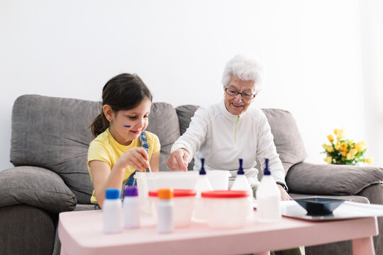 Smiling granny in eyeglasses helping positive granddaughter with dirty face in casual wear preparing blend using colorants in plastic container while sitting on comfortable couch in living room