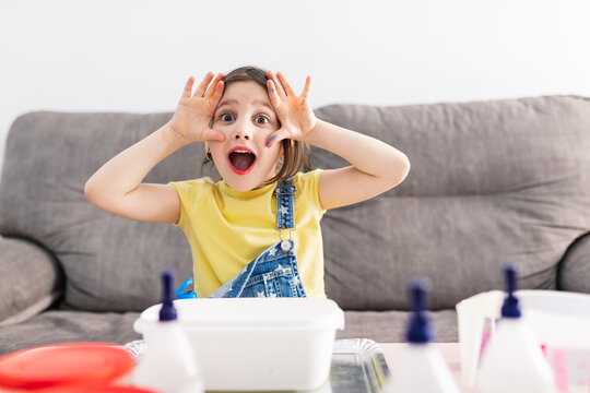 Charming little child in denim overall grimacing while sitting on cozy couch with open mouth and looking at camera in front of table with plastic container and bottles