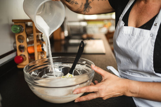 Crop housewife wearing apron standing at counter in kitchen and pouring sugar in glass bowl while preparing dough for pastry