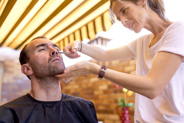 Female stylist in wristwatch trimming nose hair to bearded male client using metal scissors at home terrace