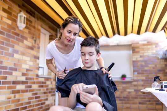 Calm boy in cape and mother with scissors and comb sharing desired haircut on cellphone on terrace