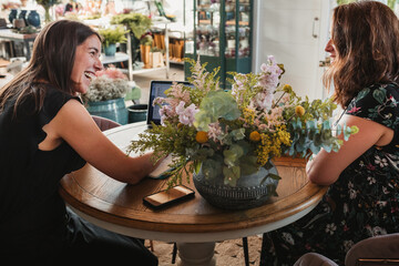Side view of female owner of stylish floristry salon discussing details of order with customer while sitting together at table with laptop in a happy attitude