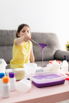 Child in yellow t shirt with spoon adding purple substance into rectangular plastic container while preparing blend and sitting on couch in living room