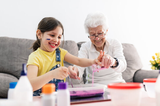 Smiling granny in eyeglasses helping positive granddaughter with dirty face in casual wear preparing blend using colorants in plastic container while sitting on comfortable couch in living room