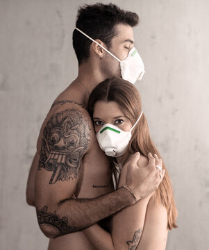 Side view of naked tattooed boyfriend and girlfriend looking at camera embracing while wearing protective masks during COVID 19 pandemic