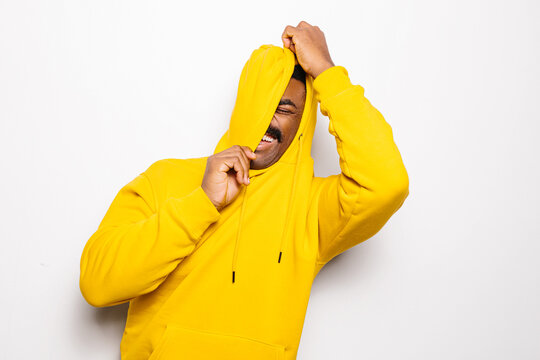 Cheerful black man over white plain background. He is looking at camera and smiling wearing a yellow hoodie. He is covering his face with the hood.