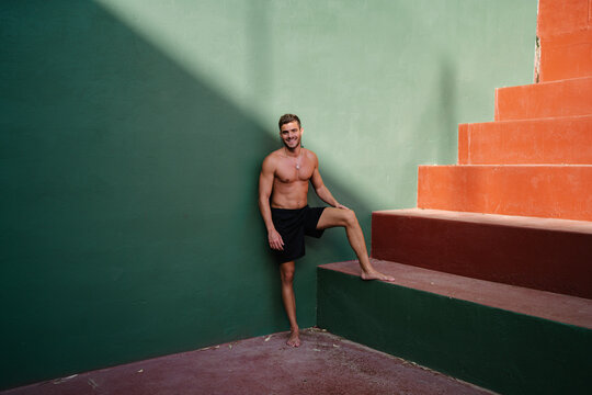 Smiling male athlete with naked torso standing barefoot near steps on sports ground and having break during intense training