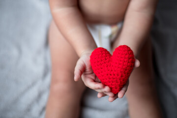 Little hands of newborn baby  with a knitted red toy heart. Health and love concept