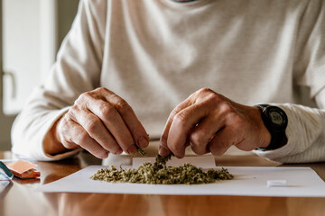 Midsection of retired elderly man preparing for rolling weed on paper at home