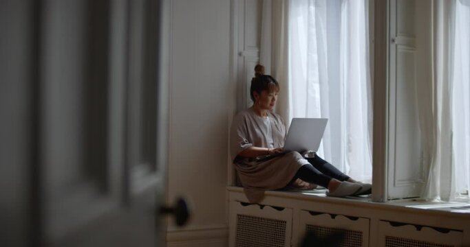 Warm sunlight peeks through window and sheer curtains as woman writes an email on her laptop while sitting on a window sill working from home
