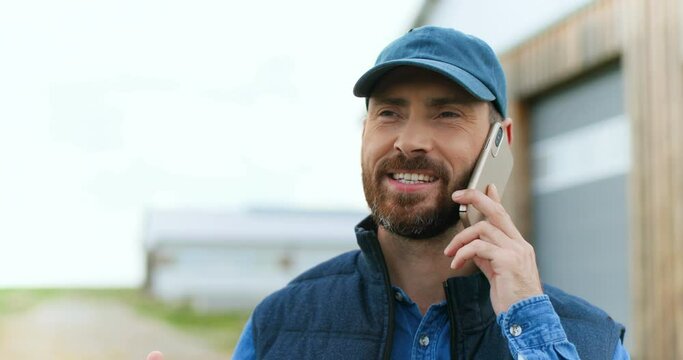 Close up of handsome cheerful Caucasian man with beard and in hat standing outdoor and talking on mobile phone at wooden shed. Happy male farmer speaking on cellphone. Telephone call. Conversation.