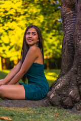 A young pretty brunette Latina with long straight hair leaning against a tree in a green dress. Portrait sitting on the grass next to a tree in the park