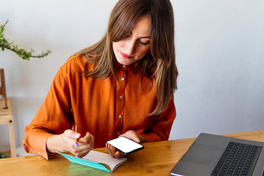 Female freelancer working at home sitting at desk with notepad and smartphone