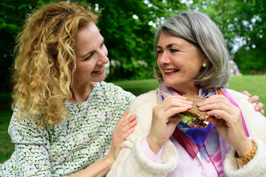 Smiling blond woman looking at senior mother eating sandwich at park