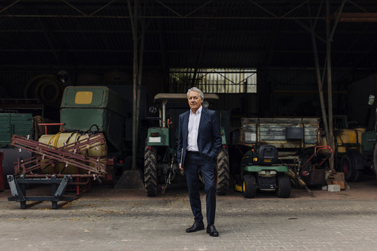Senior businessman standing on a farm with tractor in barn