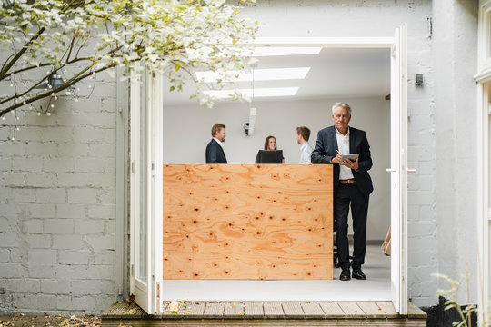 Senior businessman standing in office, using digital tablet, people working in background
