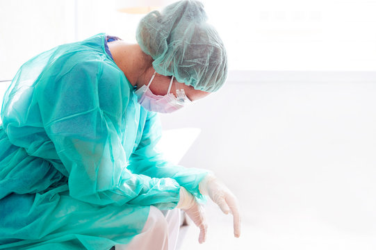 Sad female doctor wearing protective workwear sitting against window in hospital