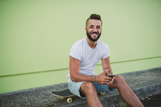 Smiling young man holding smart phone while sitting on skateboard
