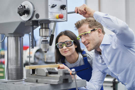 Male manager drilling metal while training female worker in metal industry