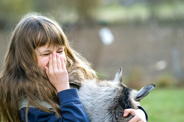 Pretty happy child girl playing with small kid goat at farm yard.