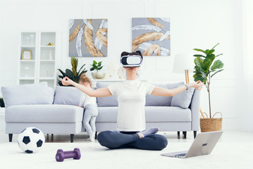 Young pretty mom meditating in lotus yoga position using virtual reality goggles while her daughter watches cartoons at home on background.