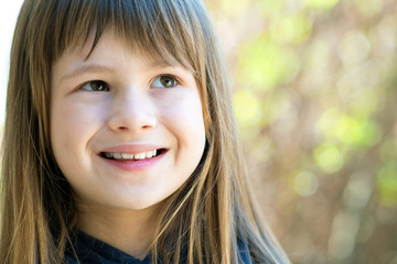 Fototapeta premium Portrait of pretty child girl with gray eyes and long fair hair smiling outdoors on blurred bright background. Cute female kid on warm summer day outside.