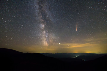 Night landscape of mountains with stars covered sky and Neowise comet with light tail.