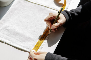 Side angle view of tailor working at table with sewing machine and marking piece of cloth with pencil while using measuring tape