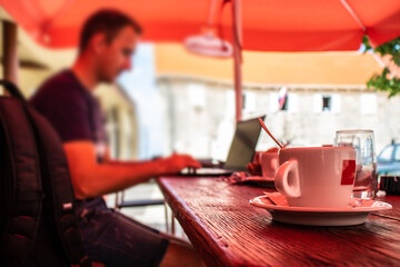 Closeup of a cup of coffee at a dalmatian coffee shop, blurred man sitting on a laptop working, concept of software developers working while travelling and being on standby