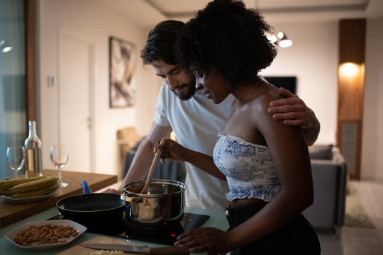 Bearded young man embracing African American woman and looking into saucepan while cooking romantic dinner in cozy kitchen together