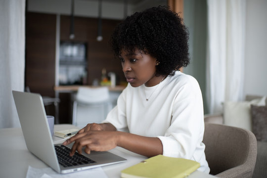 African American female with curly hair typing on laptop keyboard while sitting at table and working on remote project at home