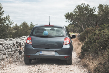 Obraz na płótnie Canvas Small grey hatchback driving on dirt roads on the island of Brac, Croatia. Driving between olive trees and rocks on small paths, throwing dust behind it. Seen from the car behind following it