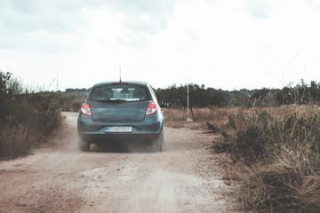 Fototapeta na wymiar Small grey hatchback driving on dirt roads on the island of Brac, Croatia. Driving between olive trees and rocks on small paths, throwing dust behind it. Seen from the car behind following it