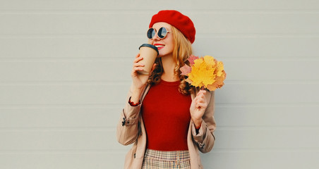 Autumn portrait of smiling young woman with yellow maple leaves and cup of coffee wearing red french beret over gray background