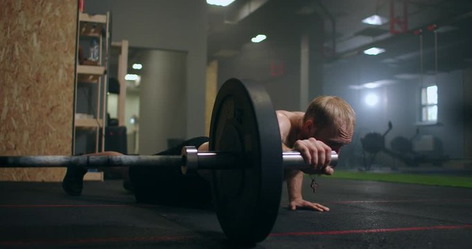 A man pushes up by rolling a barbell with one hand on the floor of the gym in slow motion
