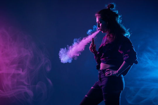 Vaper on a dark background. Young woman is vaping. Female student next to vape smoke. Woman blows steam from vape. Concept - sale of sets for vape. Concept - relax. Vapor from an electronic cigarette