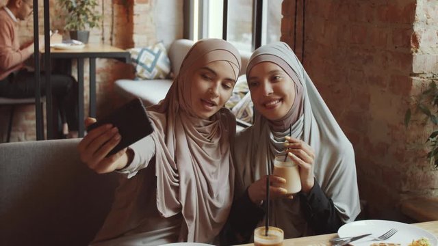 Two young muslim female friends in hijabs sitting together at cafe table, holding coffee, smiling and posing for smartphone camera while taking a selfie