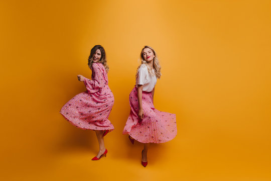 Full-length portrait of caucasian girl in red shoes dancing on yellow background. Studio photo of happy sisters in pink dresses.