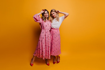 Full-length shot of attractive ladies in stylish red shoes. Good-humoured adult sisters posing on yellow background.