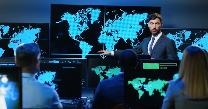 Caucasian handsome man in suit and tie having speech and explaining map on monitor. Professional male cyber security analytic demonstrating geolocation facilities on screen to colleagues.