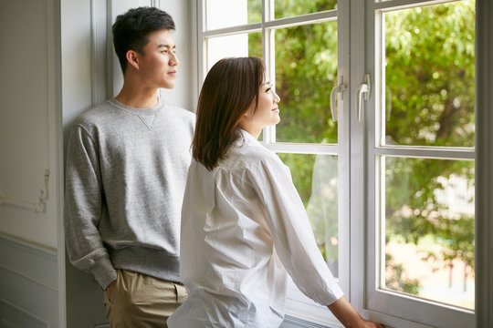 young asian couple staying at home standing by the window looking out at green trees