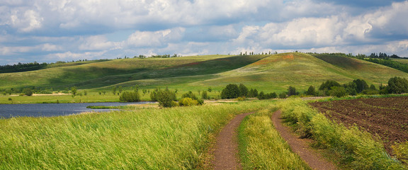 A country road among fields with grasses, near a lake and hills. Summer natural landscape with cloudy sky.