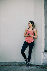 Fototapeta premium Full length of young attractive female runner leaning on the wall and using smart phone after long run in the city.